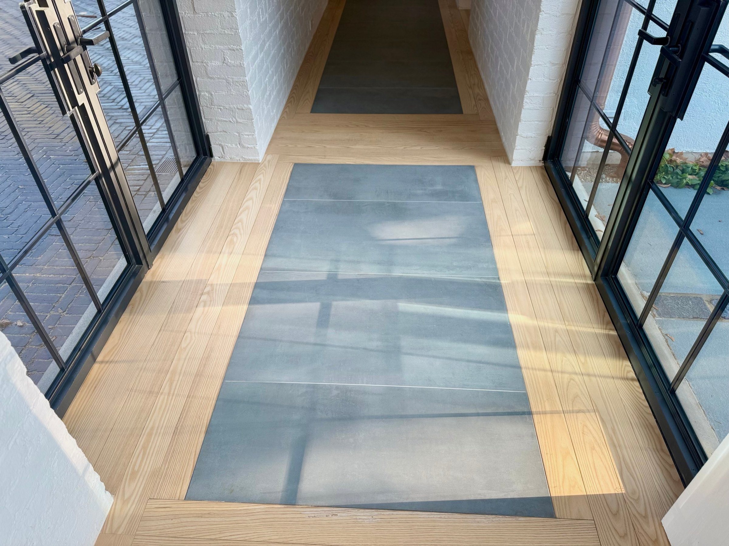Ash hardwood floor framing a panel of large-format stone at a steel-doored entry