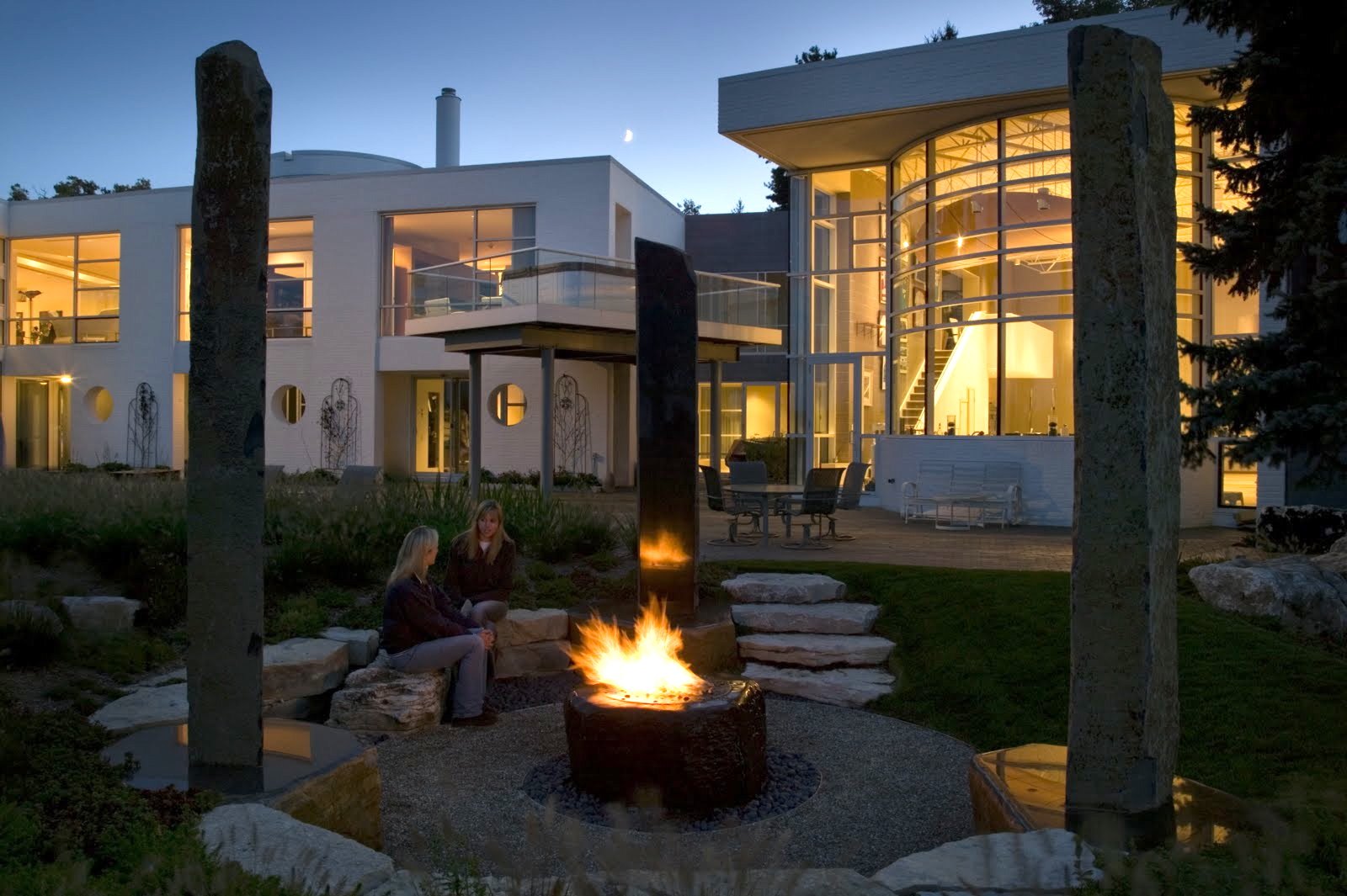 The garden at dusk: a fire pit among standing stones, with the glass-walled living room lit behind