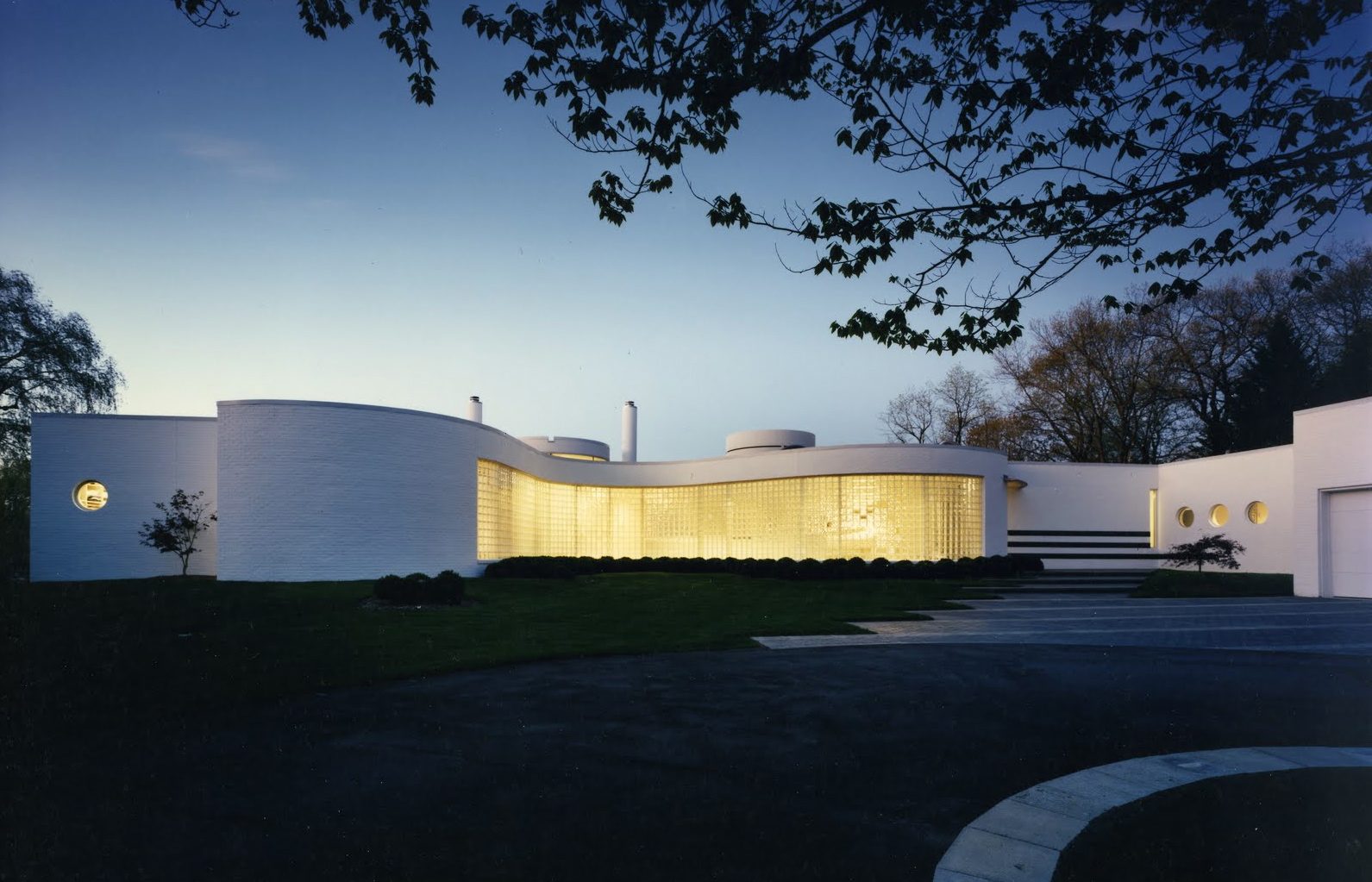 A curved white modern house at dusk, its glass-block wall glowing from within — an early Mosher Dolan home